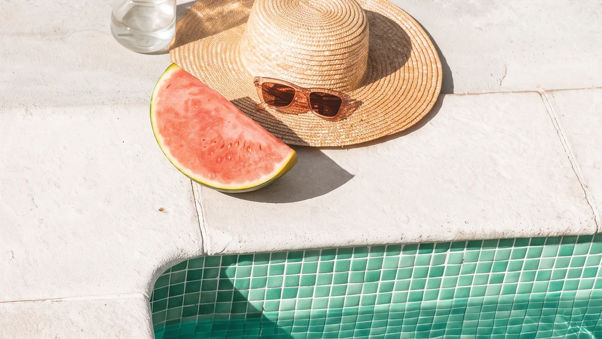 A straw hat, fresh watermelon and a glass water bottle beside sunlit jade pool water